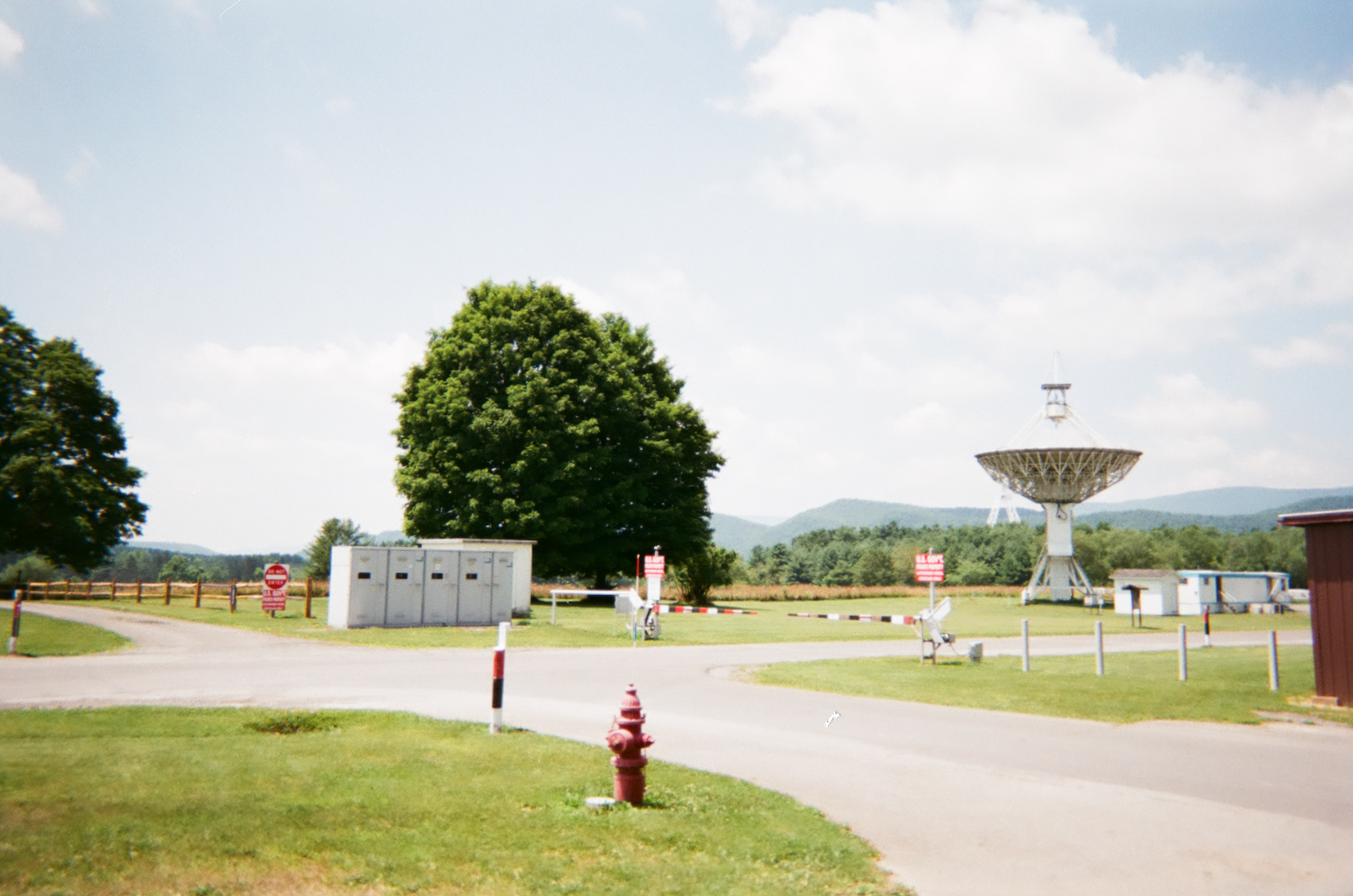 Sunny daytime photograph of a security checkpoint at the Green Bank Observatory. A red and white striped barrier arm crosses the road beside warning signs and a concrete equipment block. A large radio telescope stands to the right above rolling forested mountains. A red fire hydrant occupies the foreground.